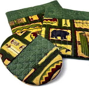 May include: A set of quilted pot holders and oven mitts. The fabric features a green background with geometric patterns, including a bear, feathers, and zigzag designs. The edges are a darker green, and the stitching is white.