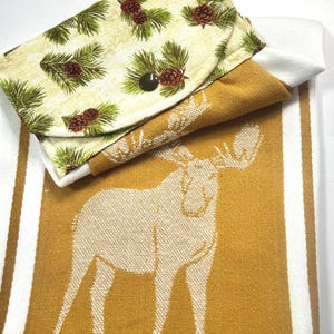 May include: A folded kitchen towel with a moose design in mustard yellow and white stripes. A small, folded pouch with a pine needle and pine cone pattern is on top. The pouch has a dark green button closure.