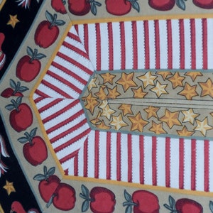May include: A fabric pattern with a repeating design of red apples, stars, and stripes. The background is a beige color with a black border.