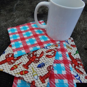 Puede incluir: Cuatro posavasos de tela a cuadros rojos y azules con un dise&ntilde;o de langosta y boya de pesca. Los posavasos est&aacute;n apilados uno encima del otro con una taza de cer&aacute;mica blanca en la parte superior.