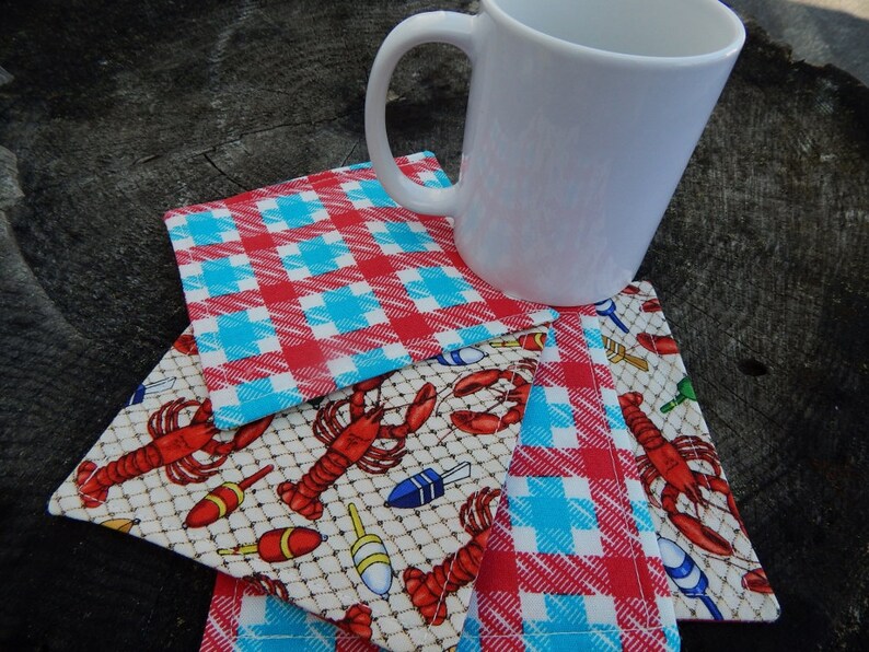 Puede incluir: Cuatro posavasos de tela a cuadros rojos y azules con un dise&ntilde;o de langosta y boya de pesca. Los posavasos est&aacute;n apilados uno encima del otro con una taza de cer&aacute;mica blanca en la parte superior.