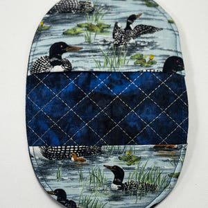 May include: An oval-shaped, quilted potholder featuring a nature scene with loons in a lake. The top and bottom sections depict the birds in a watery environment with lily pads. A dark blue, diamond-stitched fabric separates the two scenes.