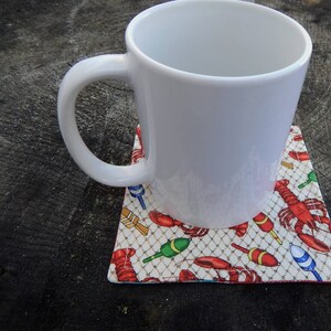 Puede incluir: Una taza de cer&aacute;mica blanca con asa sobre un posavasos con dise&ntilde;o de langosta y boya de pesca. El posavasos es blanco con detalles rojos, azules y verdes.