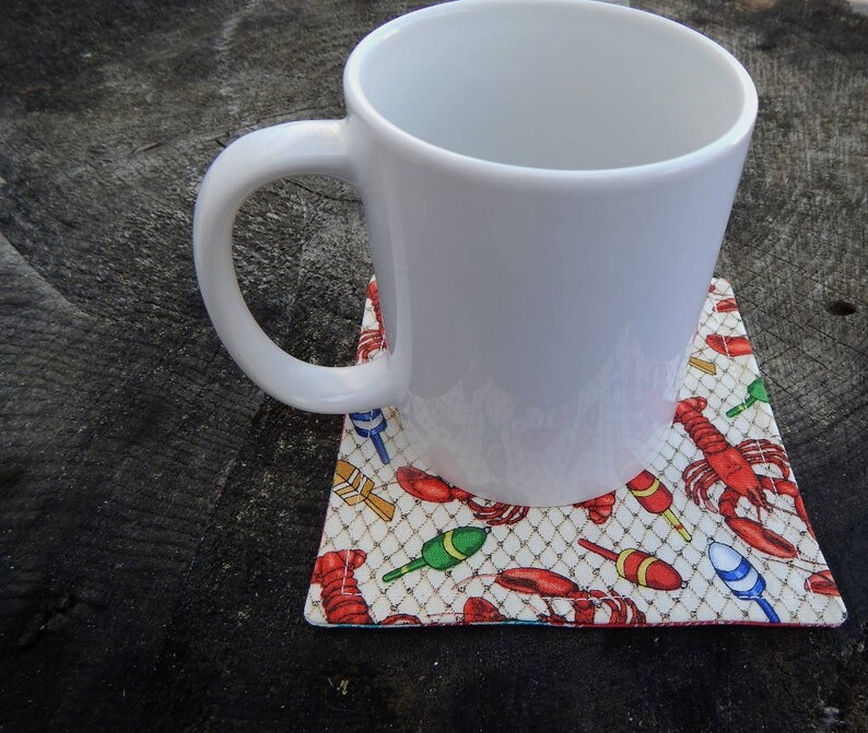 Puede incluir: Una taza de cer&aacute;mica blanca con asa sobre un posavasos con dise&ntilde;o de langosta y boya de pesca. El posavasos es blanco con detalles rojos, azules y verdes.