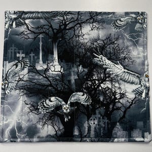 May include: A square, quilted fabric featuring a spooky graveyard scene with owls, trees, lightning, and crosses in shades of gray and white. The fabric is likely used for a placemat or similar item.