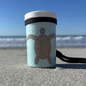 May include: A cylindrical container with a white top and a black band. The container has a light blue background with a brown sea turtle design. A black cord is attached to the side. The container is on a sandy surface with the ocean in the background.