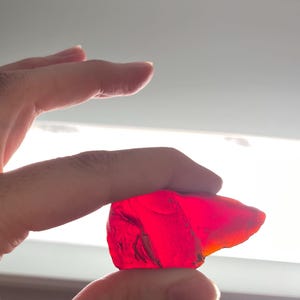 May include: A hand holding a rough, translucent, red gemstone. The stone is illuminated, showcasing its irregular shape and vibrant colour. The background is a bright, diffused light.