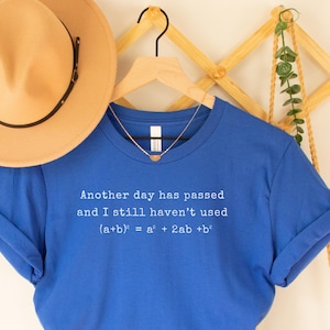 Puede incluir: Una camiseta azul real con el texto "Another day has passed and I still haven't used (a+b)² = a² + 2ab + b²". Un sombrero fedora marrón con una banda negra cuelga sobre la camiseta. También se ven una percha de madera y un bolso tejido.