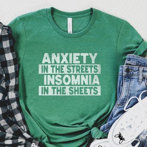 May include: A green t-shirt with the text "ANXIETY IN THE STREETS INSOMNIA IN THE SHEETS" in white. The shirt is paired with blue jeans, white sneakers, and a black and white plaid shirt.