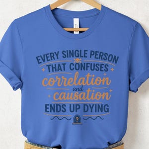 May include: A blue t-shirt with the text "EVERY SINGLE PERSON THAT CONFUSES correlation and causation ENDS UP DYING". The text is in white and orange. The shirt is paired with denim shorts and a woven belt.