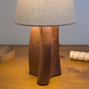 May include: A wooden table lamp with a brown base and a grey lampshade. The lamp is on a wooden table, with a wooden bowl and a wooden decorative piece. The lamp's base has a unique, sculpted design.
