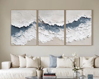 May include: Three framed abstract paintings with textured white and blue designs. The artwork features a layered, wave-like pattern with a neutral beige background. The paintings are displayed above a white sofa in a living room setting.