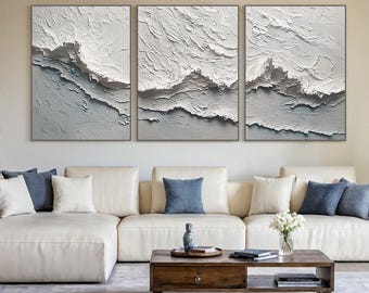 May include: Three framed abstract art pieces with textured white and gray designs. The artwork features a layered, wave-like pattern, creating a sense of depth. The pieces are displayed above a white sectional sofa with blue and white throw pillows.