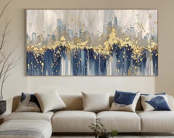 Op de afbeelding: Abstracte wandkunst met een kleurenpalet van goud, marineblauw en wit. Het kunstwerk heeft een textuur met gouden verfspatten en verticale penseelstreken. Het ingelijste schilderij hangt boven een beige hoekbank.