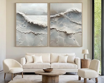 May include: Two framed abstract paintings depicting ocean waves in shades of white, gray, and beige. The textured artwork is displayed above a white sofa and chairs in a modern living room setting.