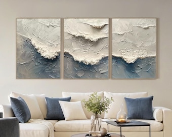May include: Three framed abstract wall art pieces. Each piece features a textured, wave-like design in shades of white, gray, and blue. The artwork is displayed above a white sectional sofa with blue and white throw pillows, creating a serene living room setting.