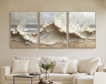 May include: Three framed abstract paintings depicting ocean waves in shades of white, beige, and gray. The textured artwork is displayed above a white sofa with matching pillows, creating a serene and modern living room aesthetic.