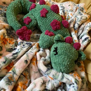 May include: A handmade, green crochet cactus with dark pink flower accents. The cactus is lying on a floral patterned blanket. The cactus has black embroidered eyes and a smiling mouth.