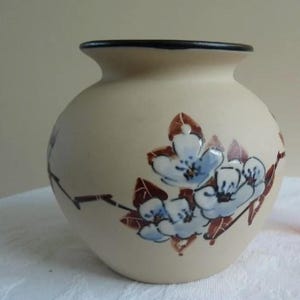 Może przedstawiać: Beżowy ceramiczny wazon o okrągłym korpusie i czarnym obramowaniu. Wazon jest ozdobiony niebiesko-białymi wzorami kwiatowymi z brązowymi liśćmi. Wazon jest przedmiotem dekoracyjnym.