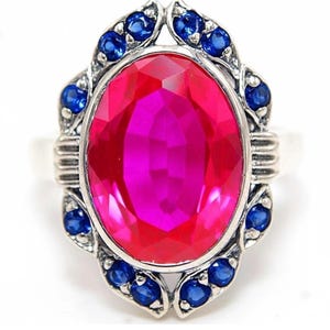 May include: A silver ring featuring a large, oval-cut, vibrant pink center stone. The central stone is surrounded by a decorative metal frame set with twelve small, round, deep blue stones. The metal band has a polished, metallic finish.