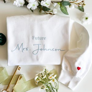 Op de afbeelding: Een wit sweatshirt met ronde hals en het geborduurde opschrift "Future Mrs. Johnson" in lichtblauw script en druklettertype. De rechtermouw bevat een klein, rood geborduurd hartje bij de geribbelde boord op de witte stof.