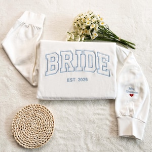 Könnte beinhalten: Weißer Pullover mit blau gesticktem Text "BRIDE EST 2025" und einem kleinen roten Herz mit dem Text "Mrs. Strongman" auf dem Ärmel.