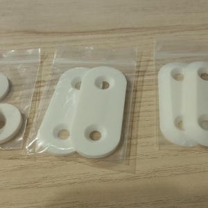 May include: Several small plastic bags containing white hardware components. The bags hold round washers and rectangular pieces with two holes each. The items are arranged on a light wood surface.
