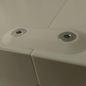 May include: A close-up view of a white furniture corner joint, featuring two metal screws and washers. These are set into a white, oval-shaped plastic piece, connecting two white panels. The image focuses on the hardware and construction details.