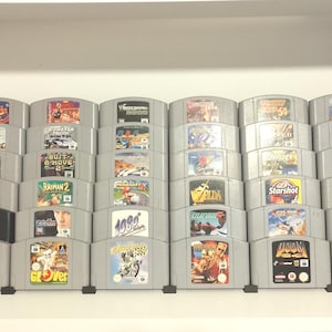 May include: A collection of thirty-one Nintendo 64 video game cartridges arranged in six rows on a shelf. The cartridges are primarily grey with one black cartridge, each featuring a unique colourful label with game titles and graphic artwork.