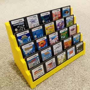 May include: A yellow display stand holding multiple Nintendo DS game cartridges. The stand is tiered, displaying rows of game cases with colourful labels. The games include titles such as "Transformers", "Word Coach", and "Rayman".