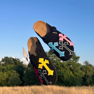 May include: Black suede clogs featuring cross designs in pink, blue, yellow, and red. The clogs have a cork footbed and adjustable straps. The shoes are displayed against a backdrop of a blue sky and green trees.