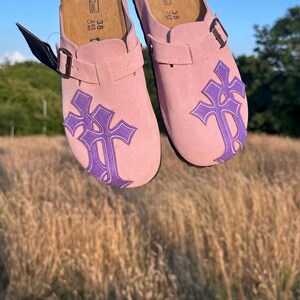 May include: Pink suede clogs featuring a purple cross design. These clogs have adjustable buckles and cork footbeds. The size is 38, equivalent to a UK size 5. The clogs are set against a backdrop of a field and blue sky.