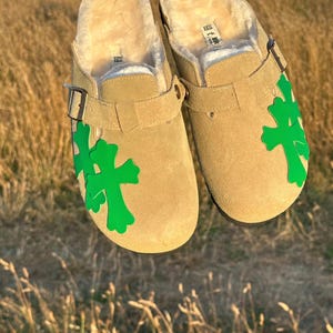 May include: Tan suede clogs with green cross detailing. The clogs feature a shearling lining and adjustable buckle straps. The shoes are displayed in a field of long grass.