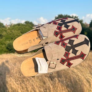 May include: Light grey suede sandals featuring a black and red cross motif. The sandals have a cork footbed, a buckle fastening, and a tag. The background shows a field of tall grass and a blue sky.