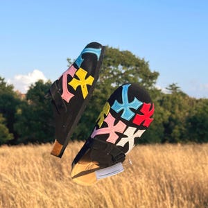 May include: Two black sandals with colourful cross-shaped designs on the footbeds are suspended in mid-air. The sandals have black straps and cork soles. The background features a field of tall grass, trees, and a blue sky.
