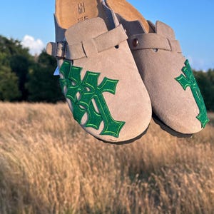 May include: Beige suede clogs with green cross-shaped detailing. These clogs feature adjustable buckles and a contoured footbed. The image shows them against a backdrop of tall grass and a blue sky.