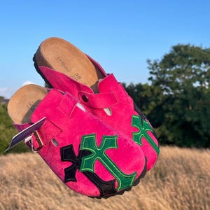 May include: Bright pink clogs featuring a green and black cross design. These clogs have a cork footbed and buckle closure. The shoes are size 38.