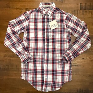 May include: A long-sleeved button-down shirt with a plaid pattern in red, white, and navy blue. The shirt has a button-down collar and a small pocket on the chest. A tag that says "THE FOX COLLECTION" is attached to the shirt.