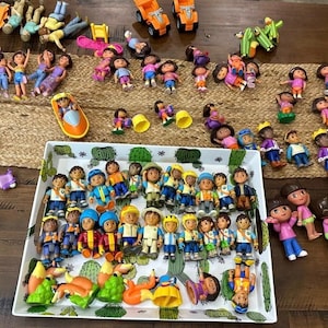 May include: A collection of Dora the Explorer figurines and toys, including dolls, vehicles, and accessories. The toys are arranged on a wooden surface and a woven mat. The dolls are in various poses and outfits, with bright colors.
