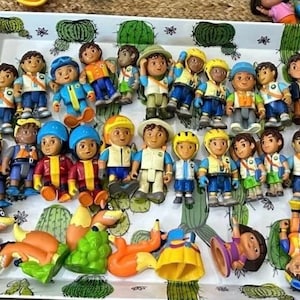 May include: A collection of colorful plastic figurines, including characters in blue, yellow, and green outfits. The figures are arranged on a white tray with a cactus pattern. Some figures are upside down.