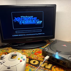 May include: A black flat-screen monitor displays the "Tokyo Xtreme Racer 2" game title. A gray Dreamcast console and a white controller with colored buttons are in the foreground. The setup is on a colorful patterned surface.