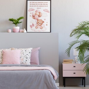 A large framed print of a teddy bear with pink balloons and Joshua 1:9 scripture displayed above a pastel-themed bed with pink and gray bedding, showing the scale of the artwork in a modern nursery room.