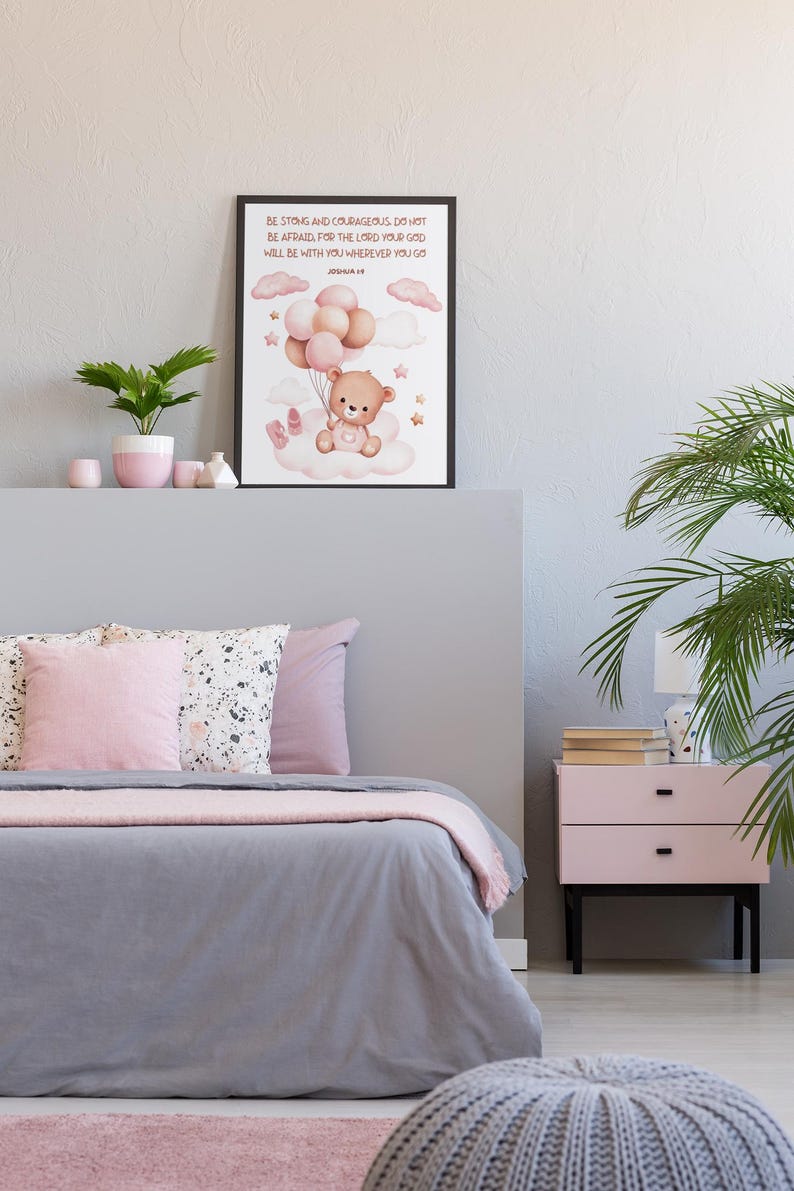 A large framed print of a teddy bear with pink balloons and Joshua 1:9 scripture displayed above a pastel-themed bed with pink and gray bedding, showing the scale of the artwork in a modern nursery room.