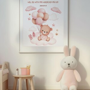 Framed wall art featuring a teddy bear with balloons and Joshua 1:9 scripture, displayed in a bright nursery with wooden furniture and a large plush bunny. The print adds a sweet and encouraging touch to the space.