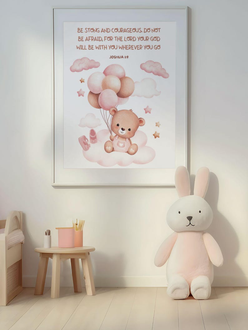 Framed wall art featuring a teddy bear with balloons and Joshua 1:9 scripture, displayed in a bright nursery with wooden furniture and a large plush bunny. The print adds a sweet and encouraging touch to the space.