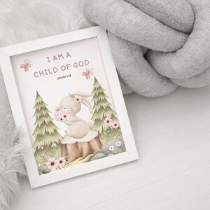 Framed bunny wall art with the verse "I Am a Child of God – John 1:12" resting on a soft white furry blanket beside a chunky gray knit cushion, styled for a gentle nursery environment.