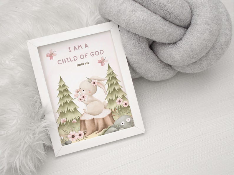 Framed bunny wall art with the verse "I Am a Child of God – John 1:12" resting on a soft white furry blanket beside a chunky gray knit cushion, styled for a gentle nursery environment.