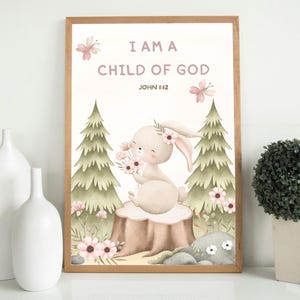 Framed nursery wall art with a bunny holding flowers and the verse "I Am a Child of God – John 1:12." Displayed on a white shelf between two white ceramic vases and a small potted plant, perfect for gender-neutral nursery decor.
