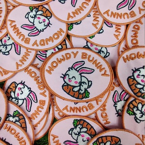 Puede incluir: Un montón de parches de tela rosa y naranja con imágenes bordadas de conejos sosteniendo zanahorias. Los parches tienen el texto "Howdy Bunny" en bordado naranja.