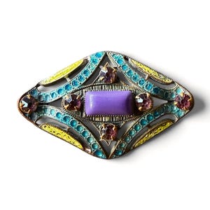 May include: A diamond-shaped brooch featuring a central rectangular purple stone surrounded by four small round purple rhinestones. The metal frame is decorated with intricate filigree patterns filled with vibrant turquoise and yellow enamel accents.